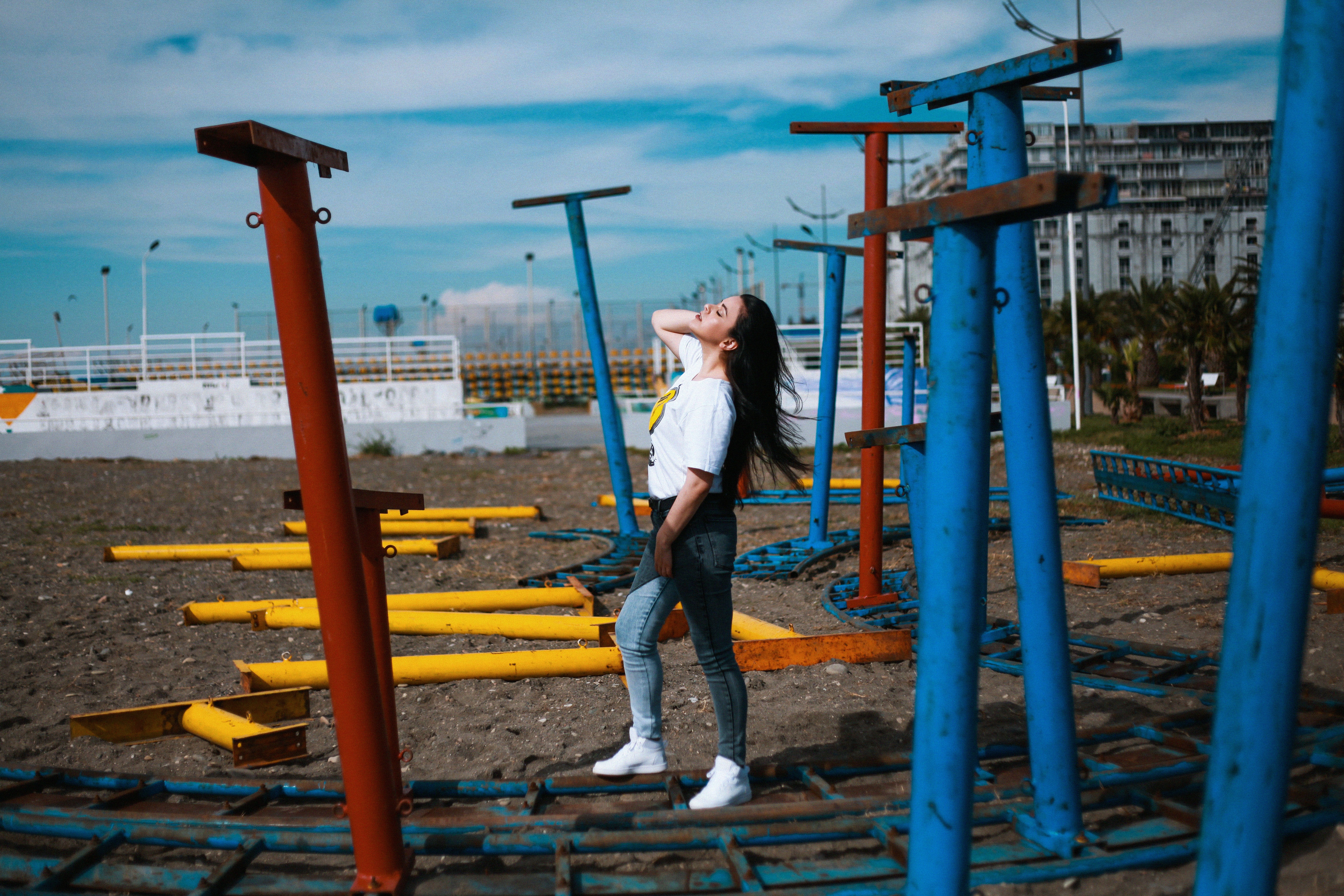 woman in white t-shirt and blue denim jeans standing in empty field beside multicolored metal stands